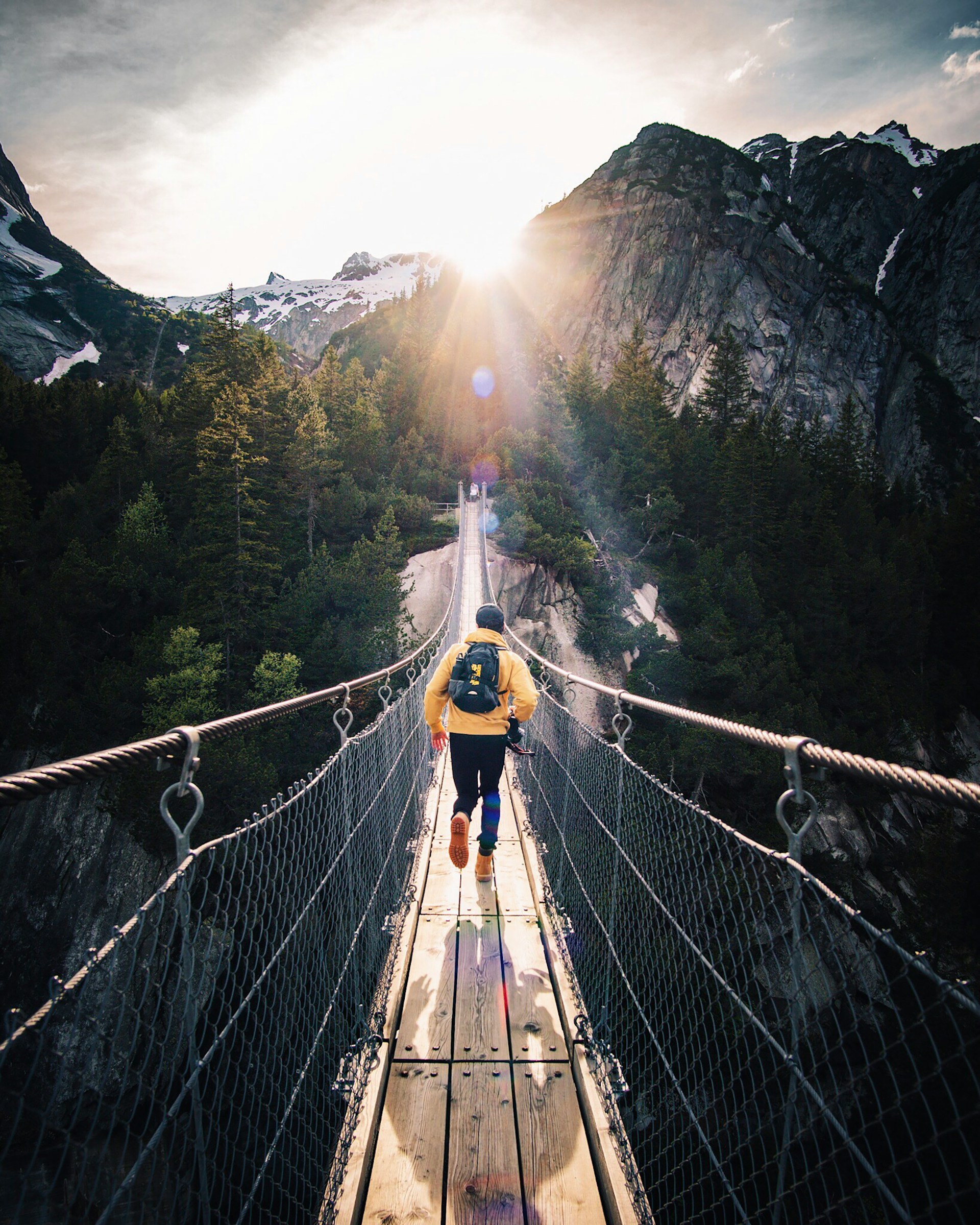 A trekker crossing a wooden suspension bridge toward sunlit mountains